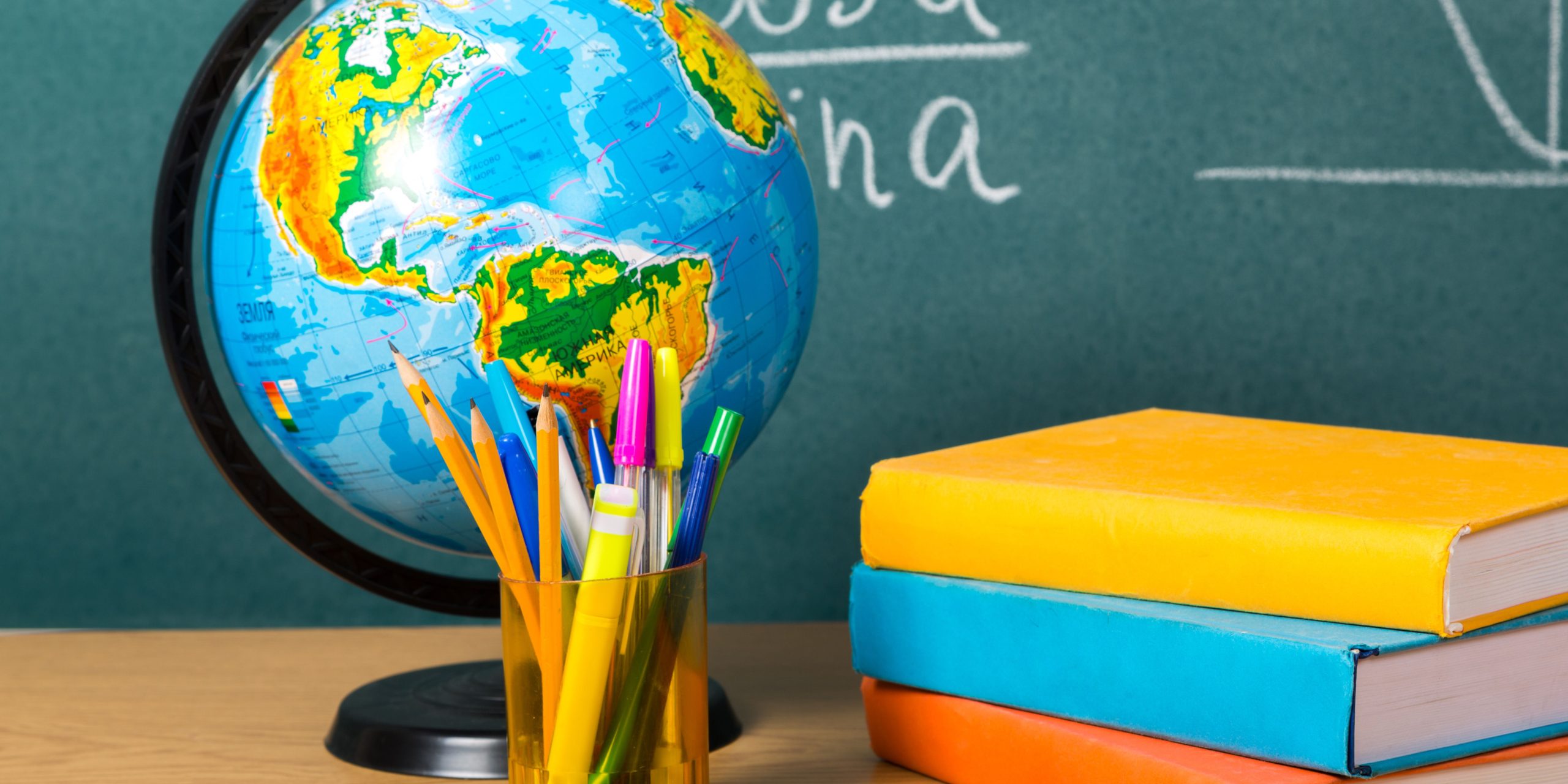 Escritorio con un lapicero, libros de colores, un globo terráqueo y de fondo una pizarra. Derechos de imagen pertenecen a Banco de Imágenes de Canva Premium.